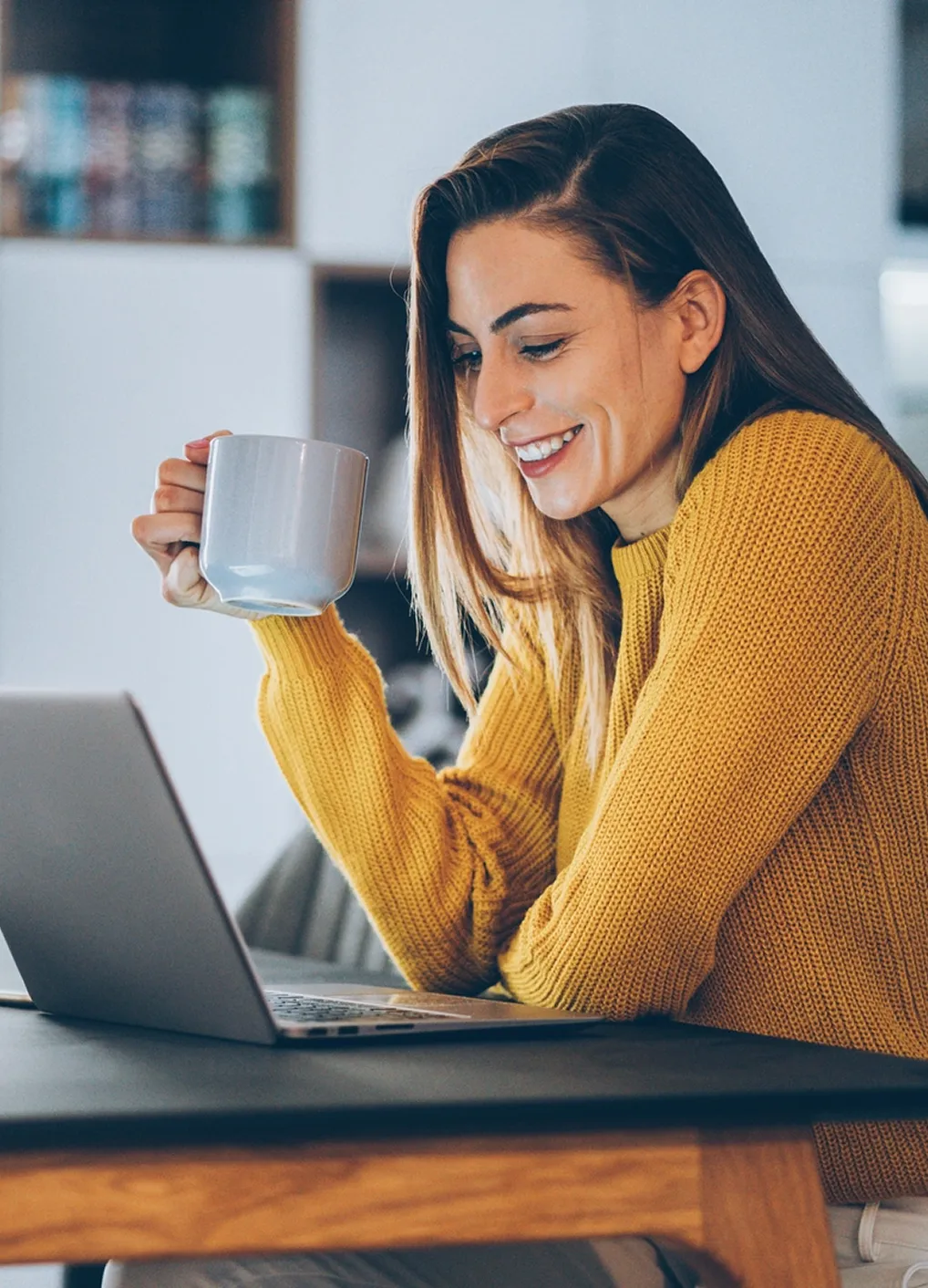 A woman smiling at her laptop while holding a coffee mug